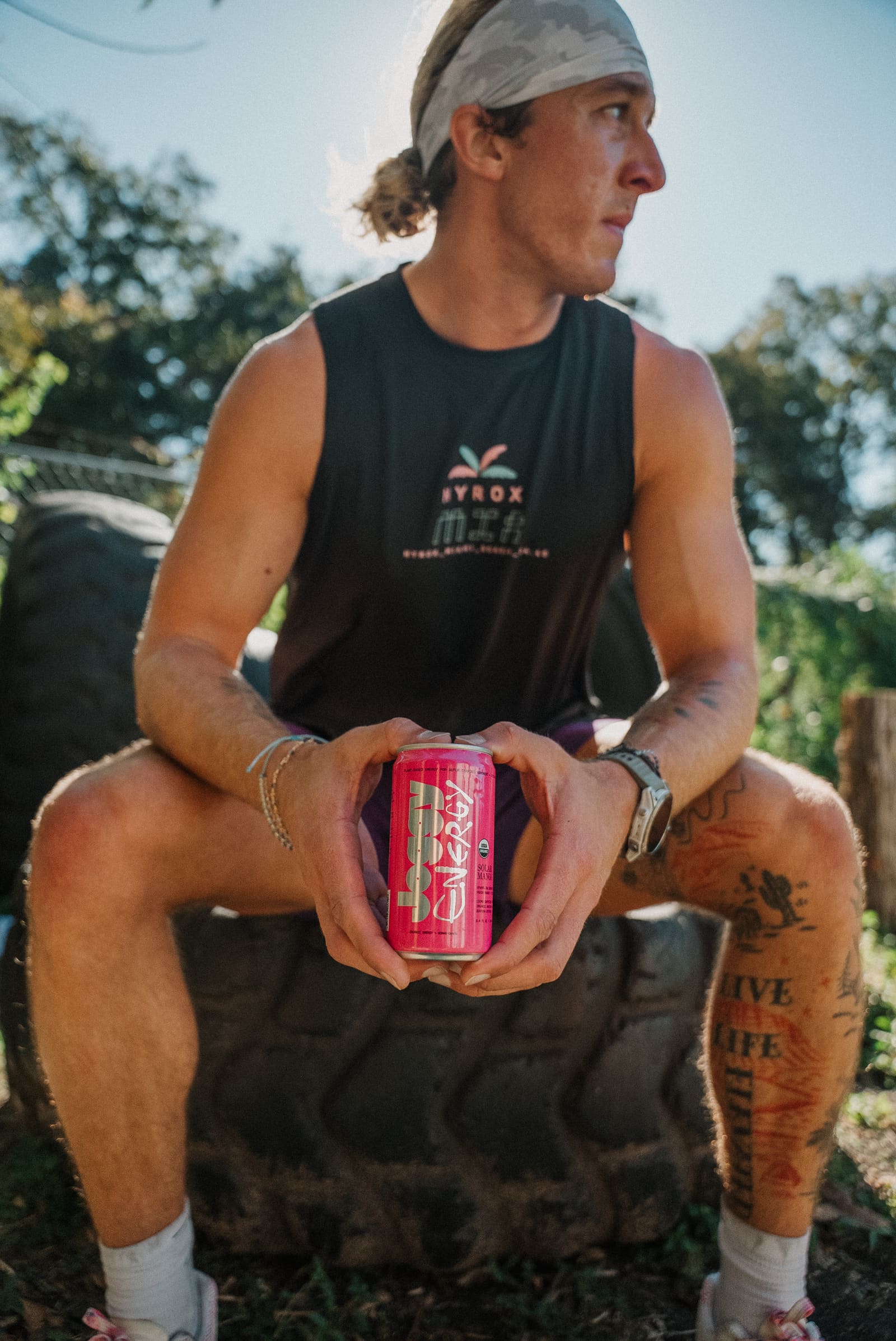 Max sitting on a training tire in a HYROX Miami tank, holding a can of Joggy Energy