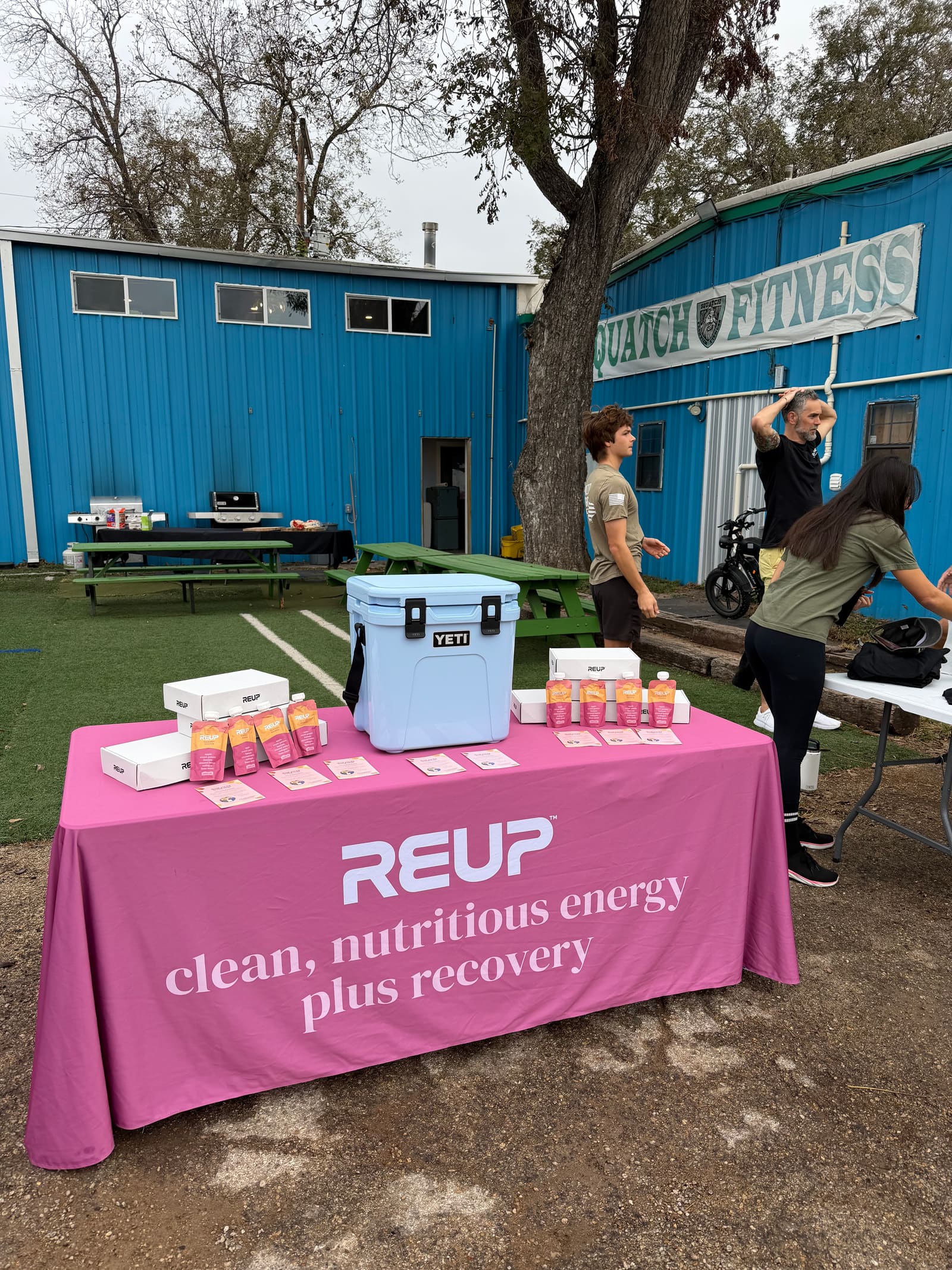 A Reup sampling table outside a gym, with a cooler and rows of smoothie pouches, while people train in the yard behind it